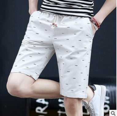 A person wearing white shorts with small black patterns and a striped shirt. The individual has their hands in the pockets and is standing against a dark background, wearing white sneakers and a red bracelet.