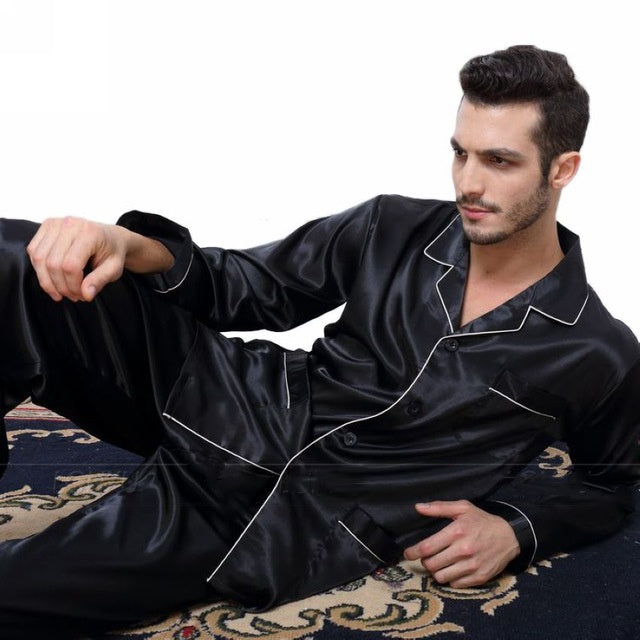 A person relaxing on a classic design rug, dressed in the Men's Satin Pajama Set - Luxurious Comfort for Lounging and Sleeping, featuring white piping. The background is plain white.