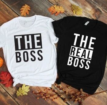 Two T-shirts from "The Boss" & "The Real Boss" Couples Matching T-Shirt Set are laid out on a wooden floor, surrounded by autumn leaves—an ideal gift for couples. The white shirt features the phrase "THE BOSS," while the black one states "THE REAL BOSS." It's a fun and playful tee set that highlights who's truly in charge!