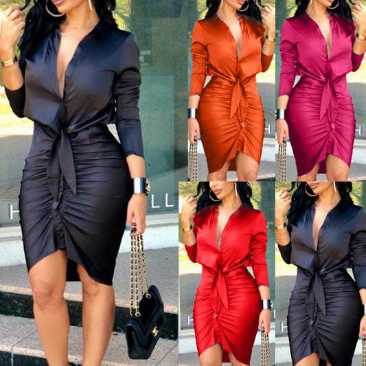 A woman showcases a chic look in the Women's Satin Ruched Bodycon Shirt Dress - Sexy Tie-Front Long Sleeve, which offers a fitted style with a front tie and ruched middle skirt. The dress is available in black, orange, burgundy, and red. Made from polyester fiber, it features long sleeves and a deep V-neckline. She completes her outfit with a black purse adorned with a gold chain strap.