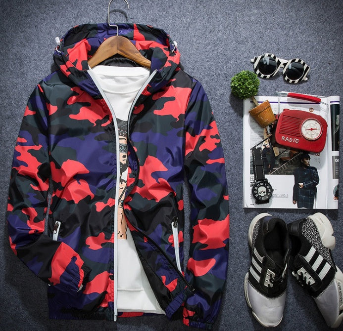 A Men's Camouflage Windbreaker Jacket, known for its lightweight and water-resistant qualities and featuring an adjustable hood, is draped over a white printed T-shirt. Nearby are black and white sneakers, sunglasses, a small potted plant, and a red vintage-style radio placed on an open magazine.