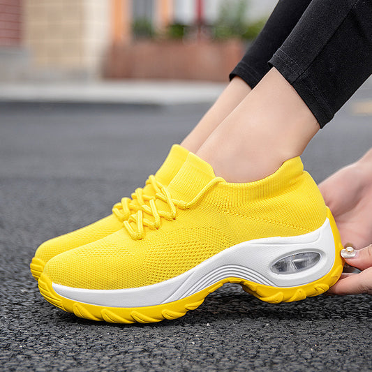 Close-up photo of a person wearing Women's Fly Knit Sock Sneakers by CJ'S, featuring a vibrant yellow color with white soles, air cushion inserts, and a flying weave design. The sneakers have matching yellow laces, and the person is adjusting the shoe on their left foot. A blurred outdoor background is visible.