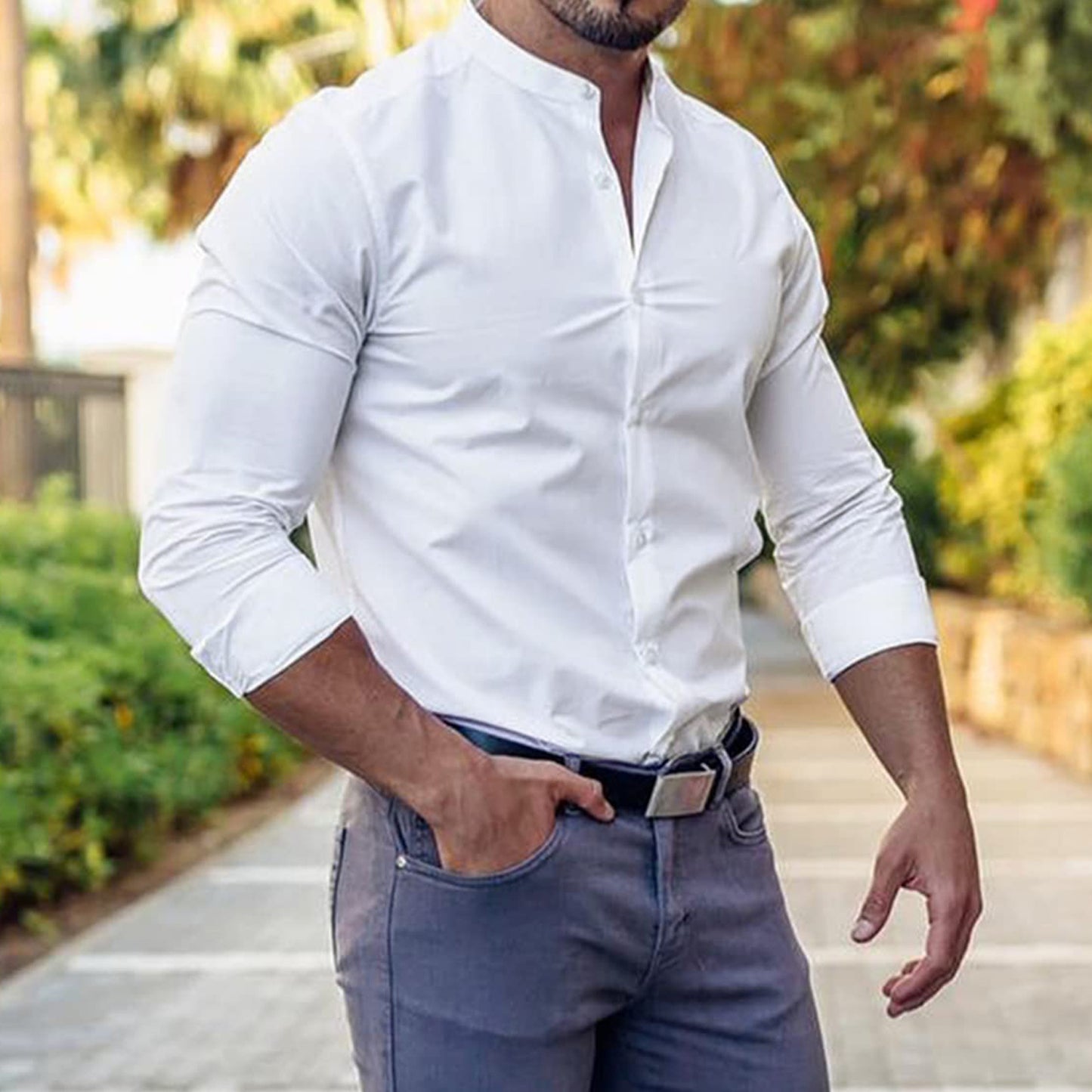 Dressed in the Men's Mandarin Collar Slim Fit Dress Shirt - Stylish and Versatile, paired with gray pants, a man stands on a pathway exuding sophistication. His left hand casually rests in his pocket, highlighted by a leather belt. The lush greenery and blurred structure in the background suggest an outdoor setting.