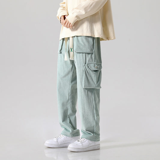 Dressed in Men's Loose All-matching Corduroy Multi-pocket Straight Cargo Pants in a light green shade, complemented by a long-sleeved cream shirt and white sneakers. The solid-color pants feature large pockets with a drawstring, set against a plain and light-colored background.