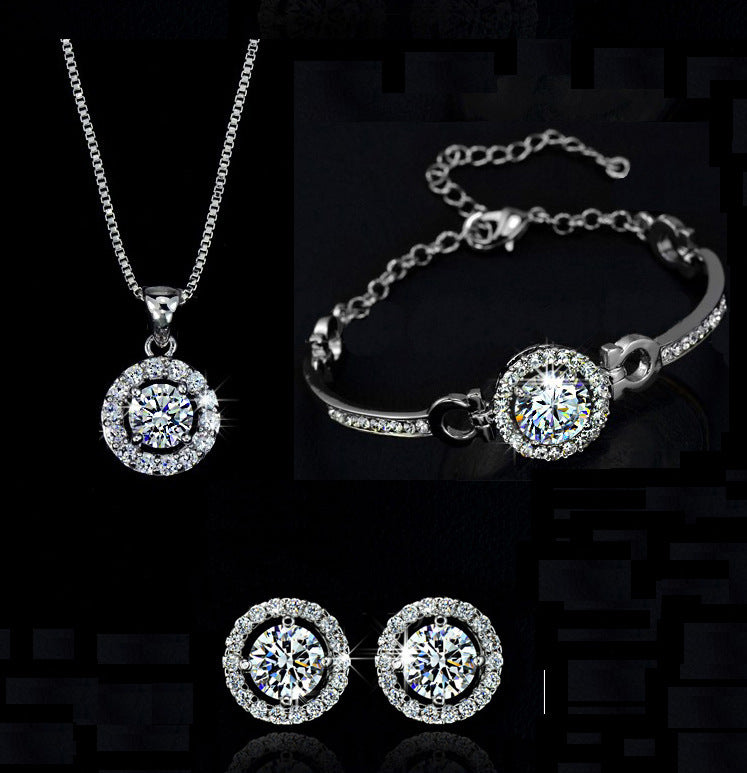 Introducing the Elegant Round-Cut Jewellery Set by CJ'S, a sparkling selection featuring a diamond-studded necklace, bracelet, and earrings. The necklace boasts a round pendant with a large central diamond. The bracelet displays a matching central diamond, while the earrings each feature a prominent diamond encircled by smaller stones.