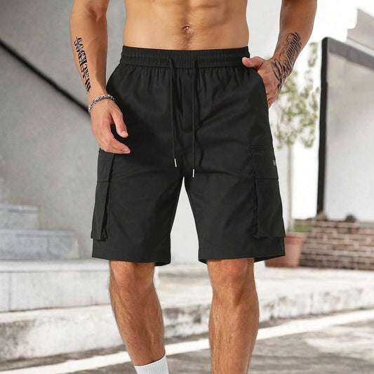 A man sporting Zendrop's Drawstring Waist Men's Multi-pocket Workwear Shorts, crafted from black polyester fiber, stands outdoors. He has tattoos on his arms and is viewed from the waist down. With a toned physique, his hands rest in the cargo pockets of the shorts. The background includes steps and a brick wall.