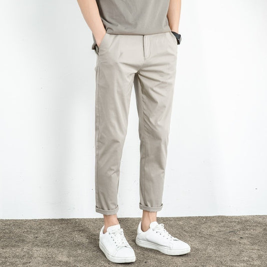 A person clad in the Men's Slim-Fit Lightweight Chino Pants, known for their comfort and style, stands atop a textured gray surface. The beige pants complement their white sneakers while their hands are casually tucked into the pockets. The versatile ensemble is highlighted against a plain white wall backdrop.