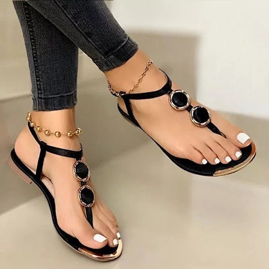 A person is wearing Women's Flat Sandals Summer Beach Shoes by CJ'S, which feature decorative gold and black circular accents made from artificial PU. The sandals are paired with an anklet adorned with small gold beads. They have white nail polish on their toenails and are styled in dark jeans rolled up to the ankles, embodying a chic Europe and America look.