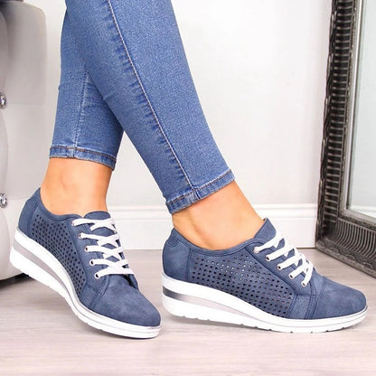 A woman wearing blue jeans and CJ'S Women's Breathable Canvas Wedge Sneakers in navy blue with white laces and white soles sits with crossed legs on a light-colored floor. A mirror and part of a piece of furniture are visible in the background.