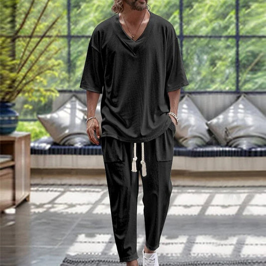 A person with long hair and a beard is dressed casually in the CJ'S Men's Casual Solid Colour Pullover V-neck Short Tube Drawstring Cufflinks Design Trousers Suit in black. They are wearing white sneakers and several bracelets, standing in a room with large windows and greenery outside.