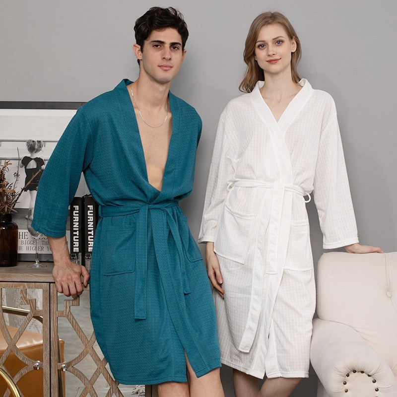 A man and a woman stand side by side wearing the Matching Couples Bathrobes - Cozy Loungewear for Men and Women, with the man in a teal robe and the woman in a white one. They are smiling in a warm, modern room featuring a decorative mirror and framed artwork in the background.