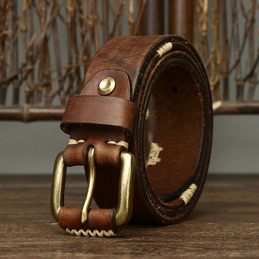 A Handmade Retro First Layer Cow Leather Belt by CJ'S with a vintage brass buckle is neatly coiled on a wooden surface. The belt features prominent stitching along its edges and a single pin buckle. In the background, vertical brown twigs enhance its rustic allure.