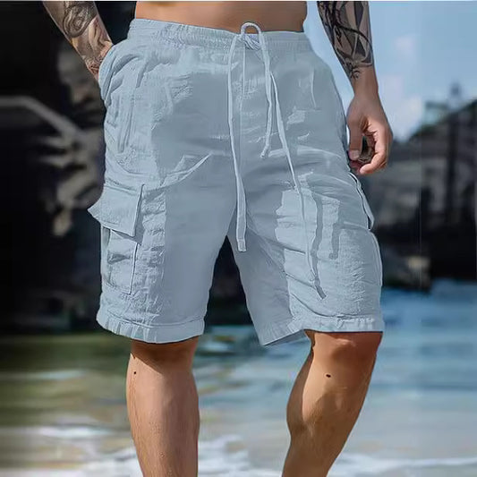 A person wearing Zendrop's Cotton Linen Shorts Drawstring Elastic Waist Straight Leg Solid Color Breathable Beach Shorts in light grey, with hands in the multiple pockets. The shorts feature a drawstring at the waist. The person has tattoos on their arms and is standing on a sandy beach with the ocean blurred in the background.