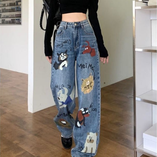 A person is wearing Zendrop's Female American retro high street graffiti jeans, which are high-waisted blue denim trousers adorned with various cartoon animal drawings. They have paired it with a black crop top and black shoes. The scene seems to be an indoor room featuring wooden flooring and shelves in the background, evoking a Japanese and Korean casual style.