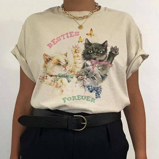 The individual is styled in street fashion, wearing a Cute Kitten Besties Forever Women's Graphic Top by Zendrop. The beige cotton t-shirt features illustrations of three playful kittens and the phrase "Besties Forever" in vibrant lettering. The outfit is complete with a gold necklace and black pants secured with a belt.