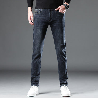 Against a gray backdrop, someone exhibits a modern style by flaunting Men's Slim-Fit Stretch Jeans paired with white sneakers. A black sweater complements the outfit, with their left hand casually tucked in their pocket and a black watch enhancing the contemporary vibe of this stylish and comfortable denim.