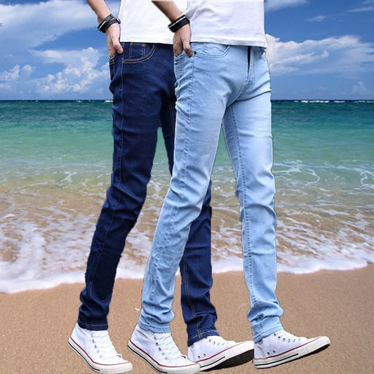 Two individuals dressed in Men's Slim Fit Stretch Jeans and white sneakers walk along a sandy beach, set against a backdrop of waves and a cloudy sky, their faces out of view.