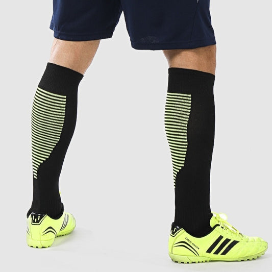 A person wearing Zendrop's Thin Soccer Socks Men's Breathable Training Sports Children's Striped Over The Knee in black with a geometric pattern, dark blue shorts, and bright yellow and black athletic shoes stands against a light gray background.