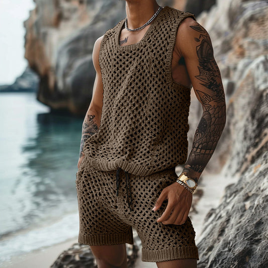 A person wearing the CJ'S Men's Lightweight Knitted Summer Set – Tank Top and Shorts stands on a rocky beach. They have tattoos on their arms and are accessorized with a necklace and a wristwatch. The background features rock formations and the ocean, making for an ideal summer fashion scene.