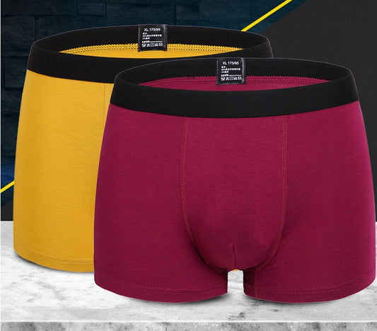 On a marble surface, two pairs of the Pure Colour Bamboo Fibre Men's Underwear are displayed, emphasizing their ultimate comfort. One pair is maroon and the other yellow, both featuring black waistbands and moisture-wicking fabric. A dark gradient background with bold yellow diagonal lines completes the scene.