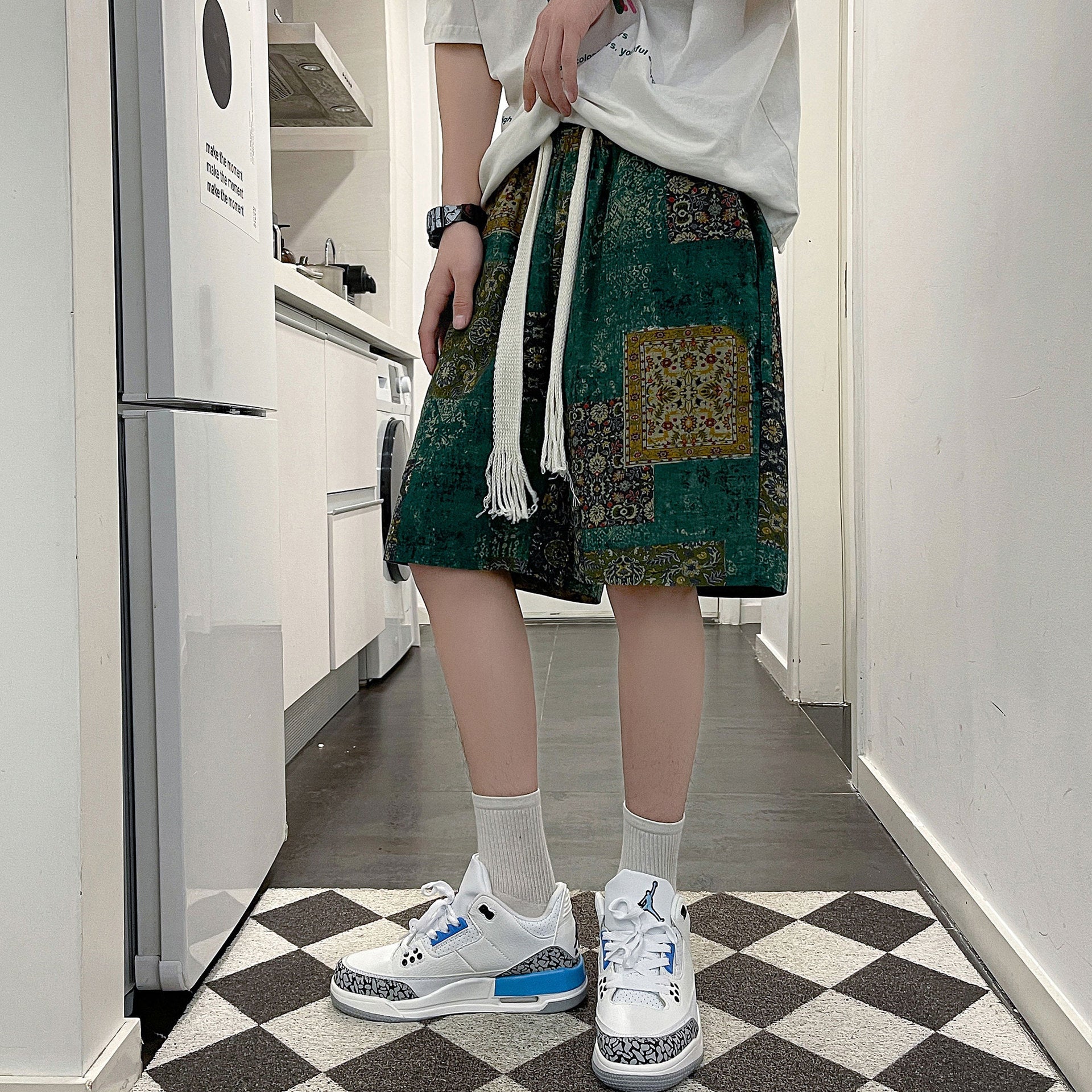 A person wearing an oversized white t-shirt, CJ's Casual Fashion Men's Sports Beach Pants featuring a cartoon pattern tied at the waist with a long white drawstring, white socks, and white sneakers with blue accents stands on a black-and-white checkered floor in a modern kitchen setting.