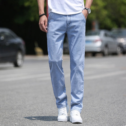 A person wearing Men's Jeans Thin Loose Straight Casual Long Pants in light blue with a non-ironing treatment and white sneakers stands on a street. Their hands are in their pockets, complemented by a white T-shirt, completing their light business look. Cars and greenery offer a bustling yet serene backdrop.