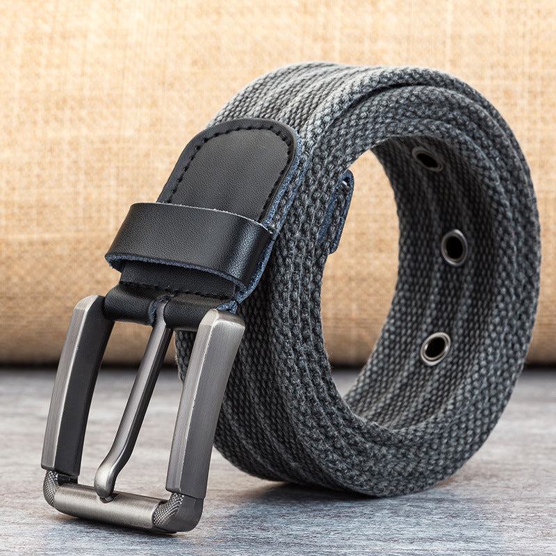 Displayed on a wooden surface is the Men's Pin Buckle Polyester Woven Pants Canvas Belt by Zendrop. The belt, featuring a gray woven fabric and black leather buckle end with a metal pin buckle, also includes silver grommet holes and is set against a beige fabric background.